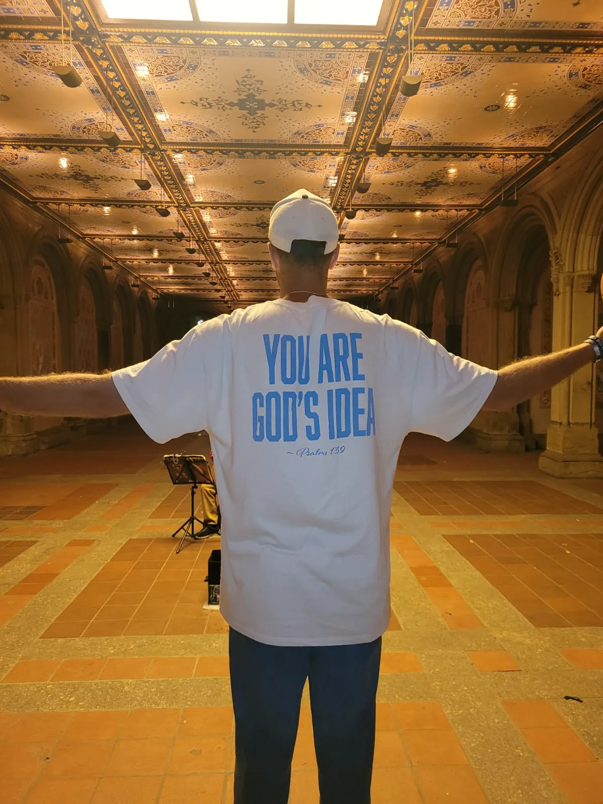 A man standing with his arms out, the back of his shirt reading You are God's idea